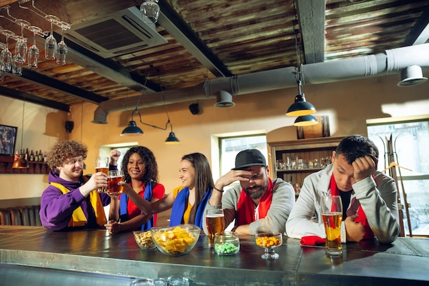 Sportfans jubeln an der Bar, im Pub und trinken Bier während der Meisterschaft, der Wettbewerb läuft. Multiethnische Freundesgruppe.