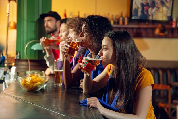 Sportfans jubeln an der Bar, im Pub und trinken Bier während der Meisterschaft, der Wettbewerb läuft. Multiethnische Freundesgruppe.