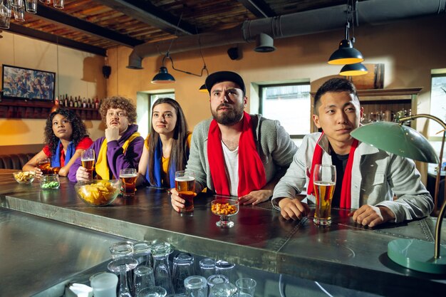 Sportfans jubeln an der Bar, im Pub und trinken Bier während der Meisterschaft, der Wettbewerb läuft. Multiethnische Gruppe von Freunden aufgeregt, die Übersetzung zu sehen. Menschliche Emotionen, Ausdruck, unterstützendes Konzept.