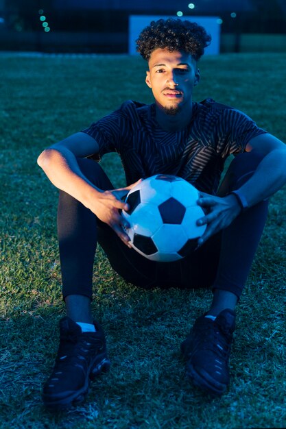 Sportler, der auf Gras sitzt und Fußball an der Dämmerung hält