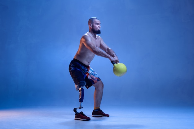 Sportler mit Behinderungen oder Amputierten isoliert auf blauer Wand. Professioneller männlicher Sportler mit Beinprothesentraining mit Gewichten in Neon. Behindertensport und Überwindung, Wellnesskonzept.