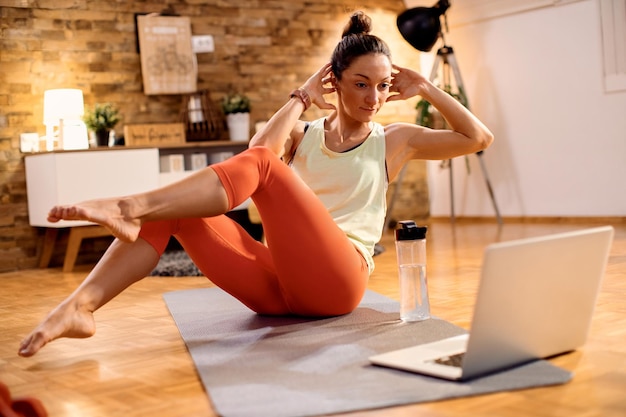 Sportlerin, die Bauchmuskelübungen macht, während sie zu Hause dem Online-Tutorial über den Laptop folgt