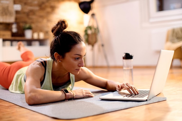 Sportlerin mittleren Alters benutzt Computer, während sie zu Hause auf dem Boden übt