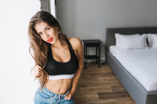Sportlich attraktives Modell in schwarzem T-Shirt, Blue Jeans entspannend in moderner Wohnung. Modisches Mädchen mit langen brünetten Haaren, roten Lippen zu Hause. Sportlicher gesunder Lebensstil, wahre Emotion.