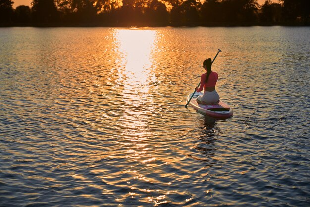 Sportliche Frau, die den Sonnenuntergang über dem See beim Sup-Boarding genießt