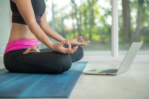 Sportliche Frau, die Yoga praktiziert, Ardha Padmasana-Übungen macht, in Lotuspose meditiert, trainiert, Sportkleidung trägt, Fitness-Video-Tutorials online auf dem Laptop ansieht, zu Hause trainieren