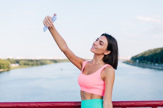 Sportliche Frau in passender Sportkleidung bei Sonnenuntergang an der modernen Brücke mit Blick auf den Fluss glückliches positives Lächeln mit Handy machen Foto-Selfie-Video für Socials-Geschichten