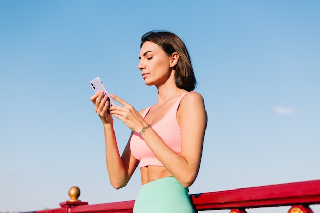 Sportliche Frau in passender Sportkleidung bei Sonnenuntergang an der modernen Brücke mit Flussblick glückliches positives Lächeln mit Handyblick auf Bildschirm