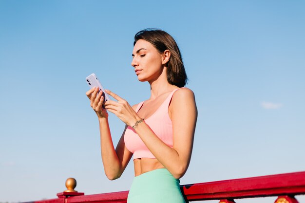 Sportliche Frau in passender Sportkleidung bei Sonnenuntergang an der modernen Brücke mit Flussblick glückliches positives Lächeln mit Handyblick auf Bildschirm