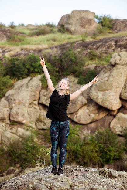 Sportliche Frau lächelnd, Frieden zeigend, stehend auf Felsen in der Schlucht