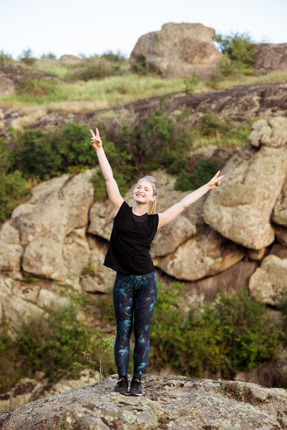 Sportliche Frau lächelnd, Frieden zeigend, stehend auf Felsen in der Schlucht