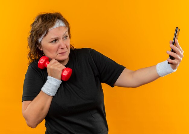 Sportliche Frau mittleren Alters im schwarzen T-Shirt mit dem Stirnband, das mit der Hantel ihres Smartphones herausarbeitet und Selfie über orange Wand steht