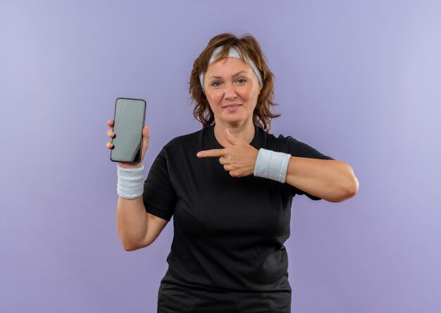 Sportliche Frau mittleren Alters im schwarzen T-Shirt mit dem Stirnband, das Smartphone zeigt, das mit dem Finger auf ihn zeigt, der zuversichtlich über der blauen Wand steht