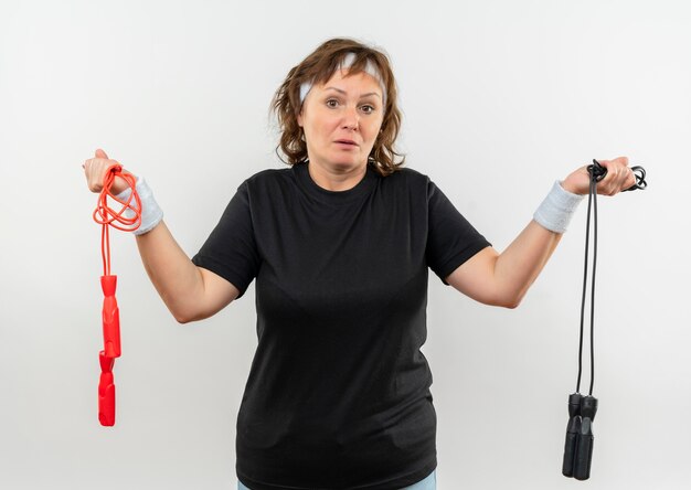 Sportliche Frau mittleren Alters im schwarzen T-Shirt mit dem Stirnband, das zwei Springseile hält, die verwirrt schauen und Zweifel haben, die über weißer Wand stehen