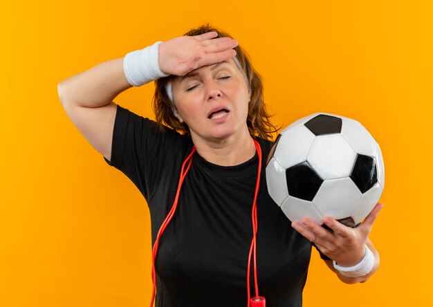 Sportliche Frau mittleren Alters im schwarzen T-Shirt mit dem Stirnband, der Fußball hält, der müde und überarbeitet steht und über orange Wand steht