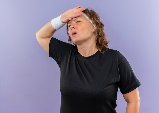 Sportliche Frau mittleren Alters im schwarzen T-Shirt mit Stirnband, das mit Hand auf Kopf müde und erschöpft nach dem Training steht, das über blauer Wand steht