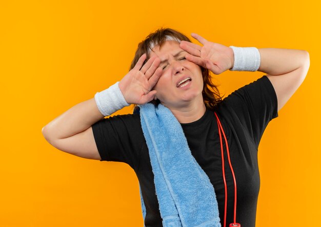 Sportliche Frau mittleren Alters im schwarzen T-Shirt mit Stirnband und Handtuch auf der Schulter, die müde und erschöpft nach dem Training über der orangefarbenen Wand stehen