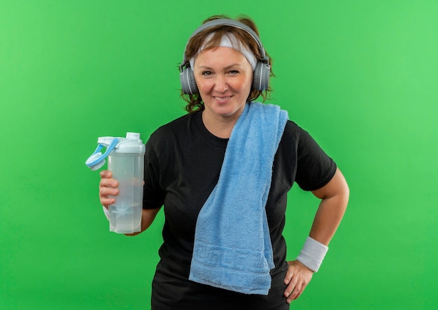 Sportliche Frau mittleren Alters im schwarzen T-Shirt mit Stirnband und Handtuch auf ihrem Hals, der Flasche Wasser lächelnd glücklich und positiv steht über grüner Wand hält