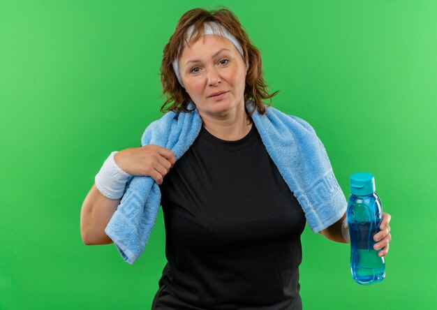 Sportliche Frau mittleren Alters im schwarzen T-Shirt mit Stirnband und Handtuch auf ihrem Hals, der Flasche Wasser verwirrt hält, das über grüner Wand steht