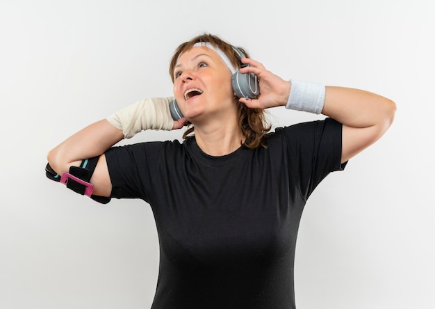 Sportliche Frau mittleren Alters im schwarzen T-Shirt mit Stirnband und Kopfhörern, die ihre Lieblingsmusik glücklich und positiv lächelnd über weißer Wand stehend genießen