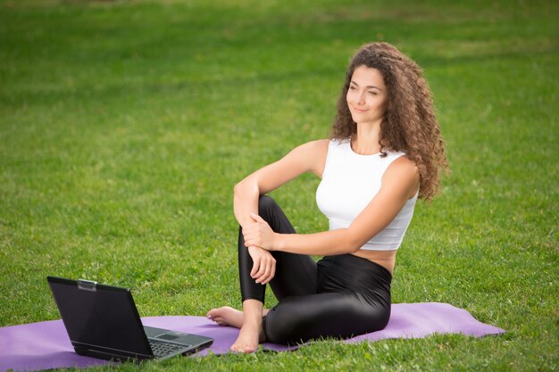 Sportliche junge Frau, die auf dem Gras mit Laptop sitzt