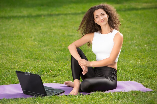 Sportliche junge Frau, die auf dem Gras mit Laptop sitzt