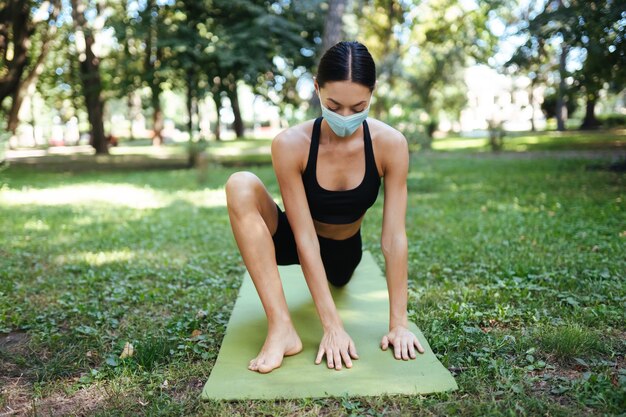 Sportliche junge Frau in einer medizinischen Schutzmaske, die am Morgen Yoga im Park macht