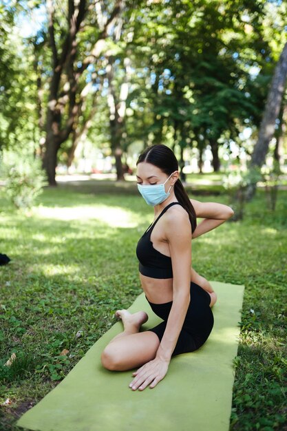 Sportliche junge Frau in einer medizinischen Schutzmaske, die morgens im Park Yoga macht, Frauentraining auf einer Yogamatte