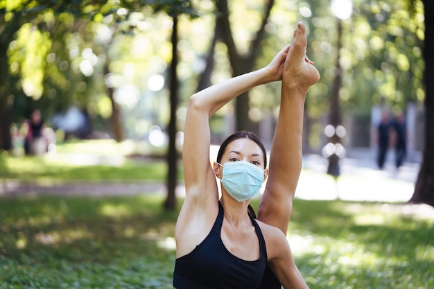 Sportliche junge Frau in einer medizinischen Schutzmaske, die morgens im Park Yoga macht, Frauentraining auf einer Yogamatte