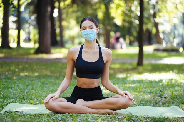 Sportliche junge Frau in einer medizinischen Schutzmaske, die morgens im Park Yoga macht, Frauentraining auf einer Yogamatte
