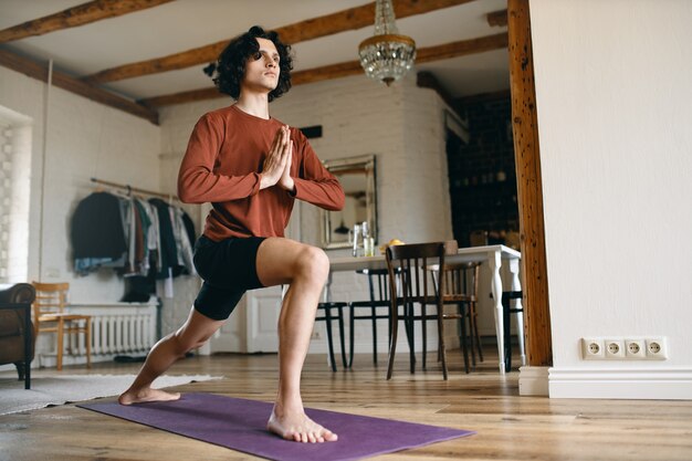 Sportlicher junger männlicher Yogi, der drinnen Yoga praktiziert, barfuß auf der Matte steht, Hände in Namaste hält und am Morgen eine Sonnengrußsequenz macht.