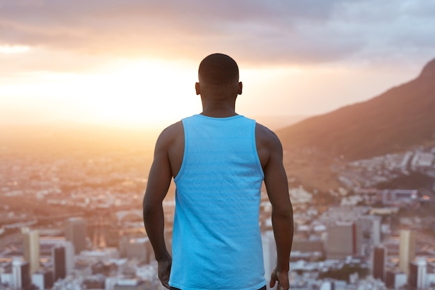 Sportlicher schwarzer Mann mit starkem muskulösem Körper, tritt zurück, denkt über etwas nach, während er den Blick auf die Natur, die Berge und die Stadt von oben bewundert und den Himmel mit Sonnenaufgang betrachtet. Sportler genießt Freiheit