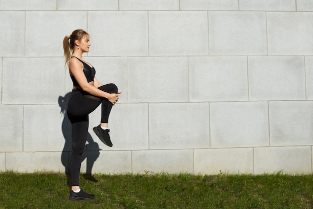 Sportliches blondes Mädchen mit Pferdeschwanz und athletischem Körpertraining im Park am Morgen, Dehnen der Beinmuskeln nach Cardio-Training, Aufwerfen gegen grauen Backsteinmauerhintergrund mit Kopienraum für Ihren Text