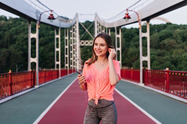 Sportliches Mädchen in den Kopfhörern, die am Stadion aufwerfen und lachen. Aktive schöne Frau, die Spaß während des Outdoor-Trainings im Sommer hat.