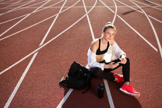 Sportliches Mädchen in drahtlosen Kopfhörern mit Rucksack und Sportflasche in der Nähe, das verträumt in die Kamera schaut, mit Handy in den Händen auf dem Laufband des Stadions