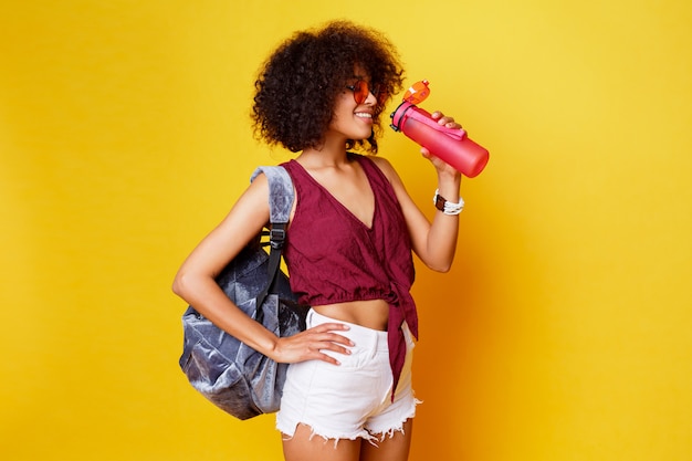 Sportschwarze Frau, die über gelbem Hintergrund steht und rosa Flasche Wasser hält. Tragen Sie stilvolle Sommerkleidung und einen Rucksack.