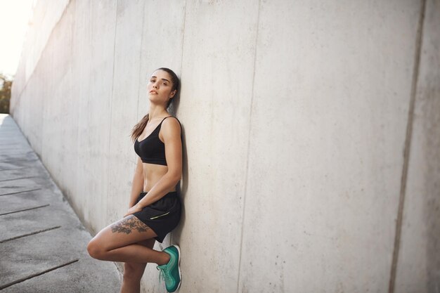 Sporttraining und gesundes aktives Lifestyle-Konzept Attraktive Sportlerin in Activewear, die nach einer produktiven Jogging-Übung eine schlanke Betonwand mit Blick auf die Kamera ruht
