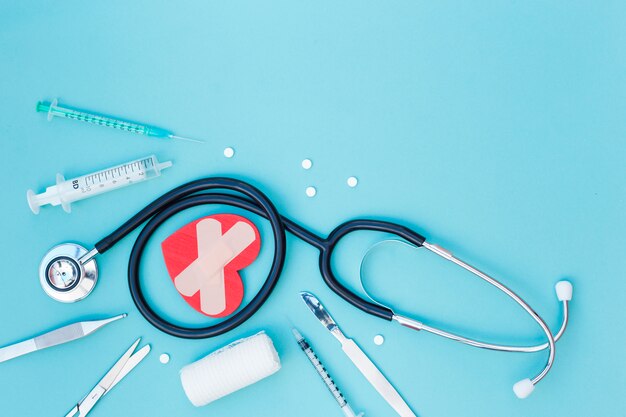 Spritze; Pillen; Stethoskop; Herzform mit Verband; medizinisches Skalpell; Schere und Baumwollgaze-Verband auf blauem Hintergrund