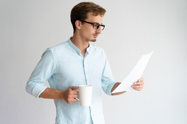 Starker hübscher junger Geschäftsmann, der Bericht analysiert und Kaffee trinkt.