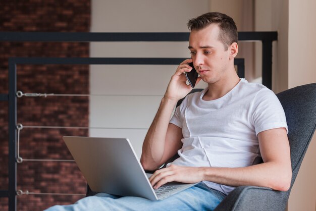 Starker Mann, der auf Smartphone spricht und auf Stuhl mit Laptop sitzt