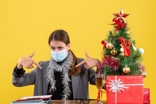 Starkes junges Mädchen der Vorderansicht mit medizinischer Maske, die am Tisch sitzt und mit Finger selbst Weihnachtsbaum und Geschenkcocktail zeigt