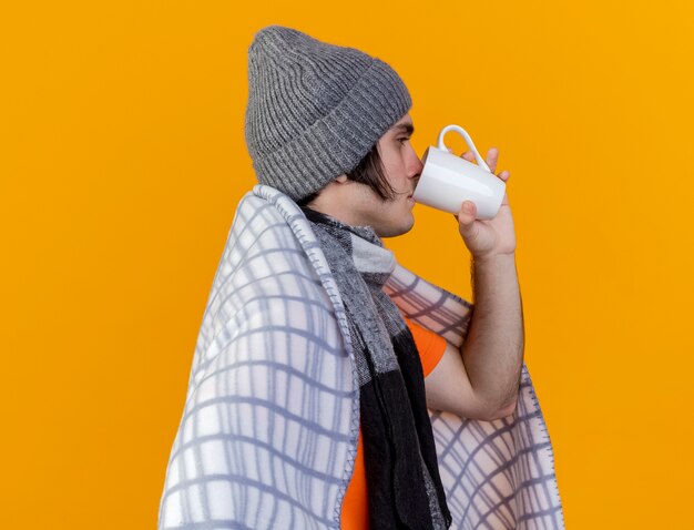 Stehen in der Profilansicht junger kranker Mann, der Wintermütze mit Schal in kariertem trinkendem Tee wickelt