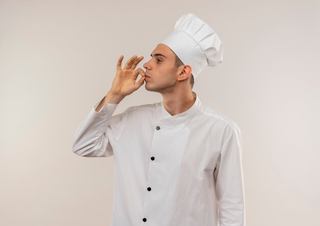 Stehen in der Profilansicht junger männlicher Koch, der Kochuniform trägt, die köstliche Geste mit Kopienraum zeigt