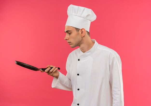 Stehen in der Profilansicht junger männlicher Koch, der Kochuniform trägt, die Pfanne in seiner Hand mit Kopienraum betrachtet