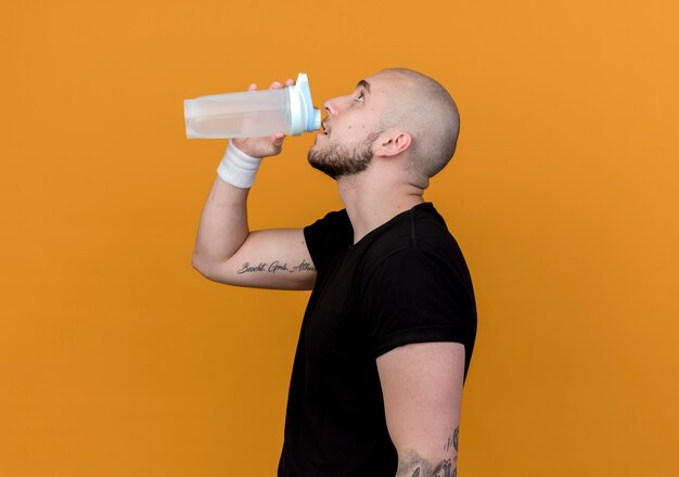 Stehen in der Profilansicht junger sportlicher Mann, der Armband trinkt, trinken Wasser von der Wasserflasche, die auf orange Wand lokalisiert wird