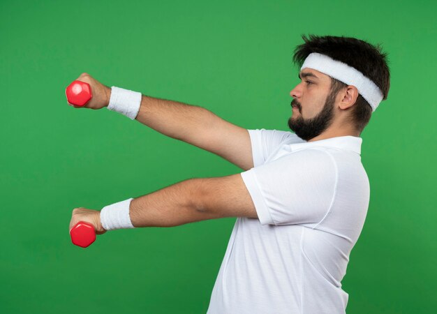 Stehen in der Profilansicht junger sportlicher Mann, der Stirnband und Armband trägt, die mit Hanteln, die auf grüner Wand lokalisiert werden