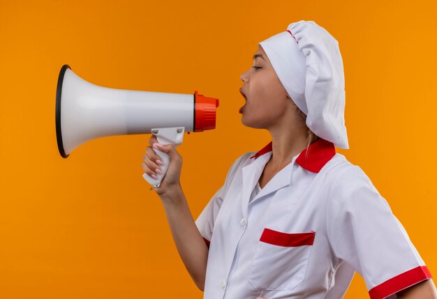 Stehend in der Profilansicht junge Kochfrau, die Kochuniform trägt, sprechen auf Lautsprecher auf lokalisiertem orange Hintergrund