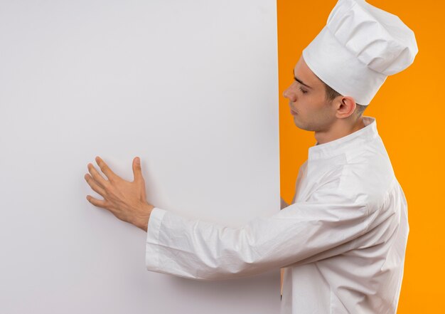 Stehend in der Profilansicht junger männlicher kühler tragender Kochuniform, die weiße Wand mit Kopienraum hält