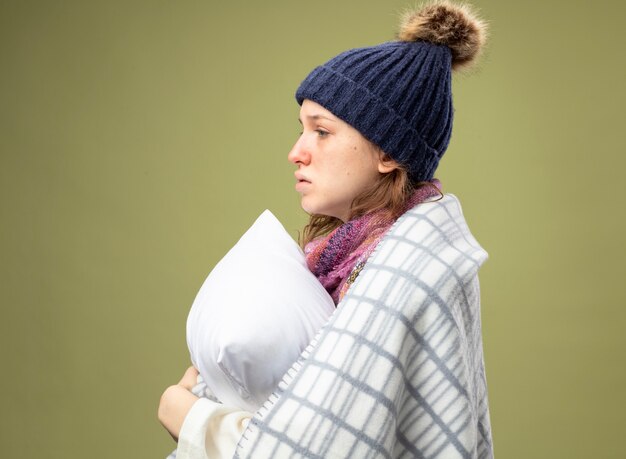 Stehend in der Profilansicht junges krankes Mädchen, das weißes Gewand und Wintermütze mit Schal wickelt, der im karierten umarmten Kissen eingewickelt wird