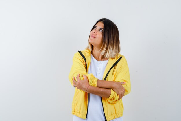 Stehende Arme des jungen Mädchens verschränkt, wegblickend im weißen T-Shirt, gelbe Jacke und fröhlich aussehend, Vorderansicht.